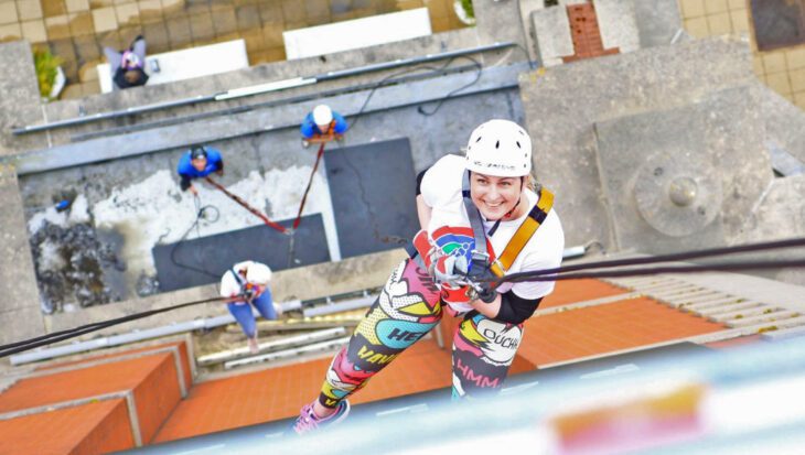 A bird's-eye view photo taken looking down from a tall tower (Trafford Palazzo) where a woman wearing a Francis House t-shirt and a helmet is making her descent and looking up to smile at the camera