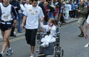 Girl in a wheelchair smiling accompanied by a runner with crowds behind