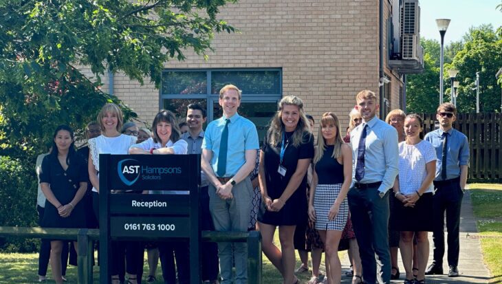 Group of people stood on the grass outside a brick office building by a sign which reads AST Hampsons Solicitors