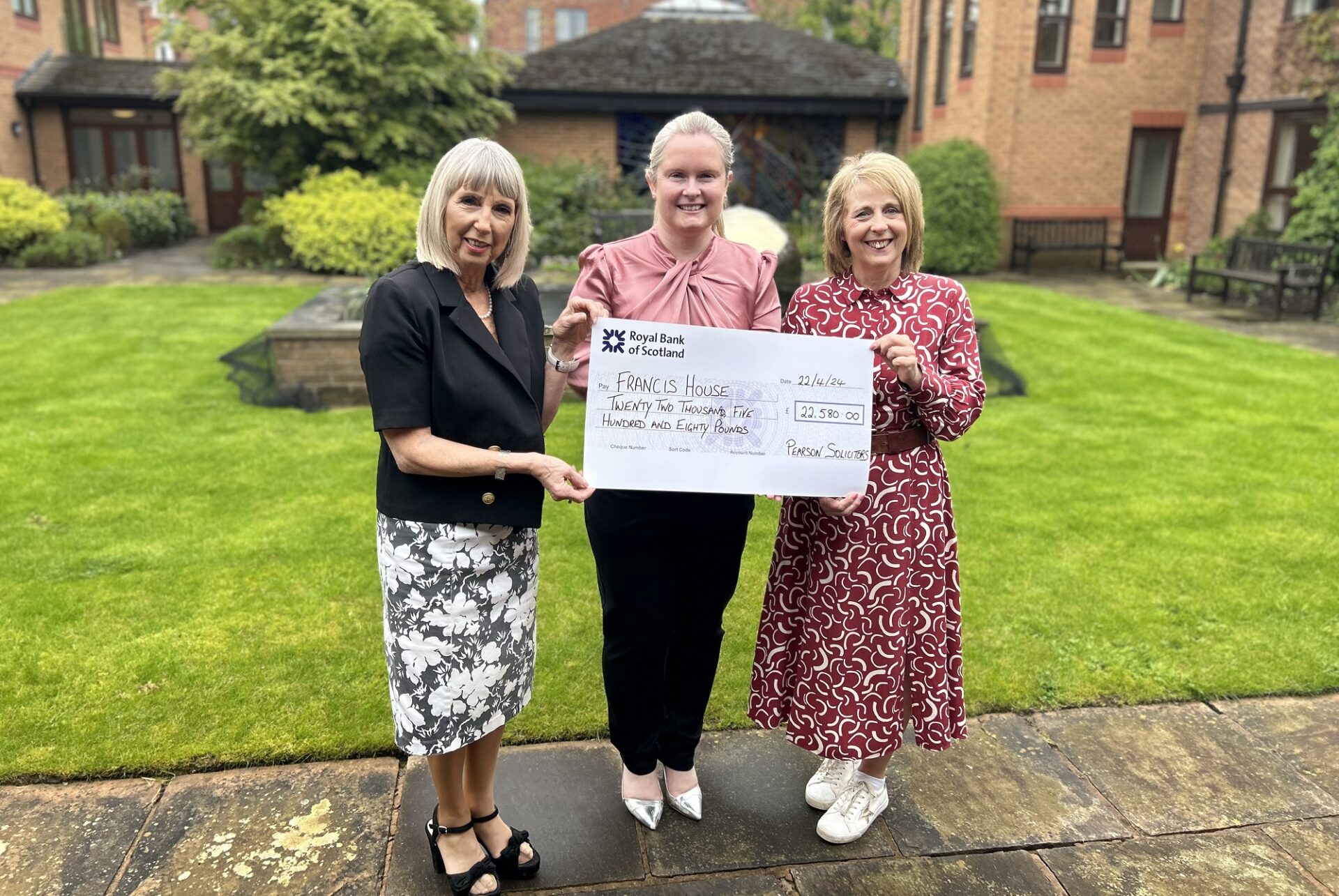 Three women stood in a garden holding a large cheque