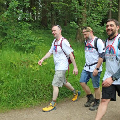 Three men wearing white t-shirts, shorts and walking boots walking along a forest path