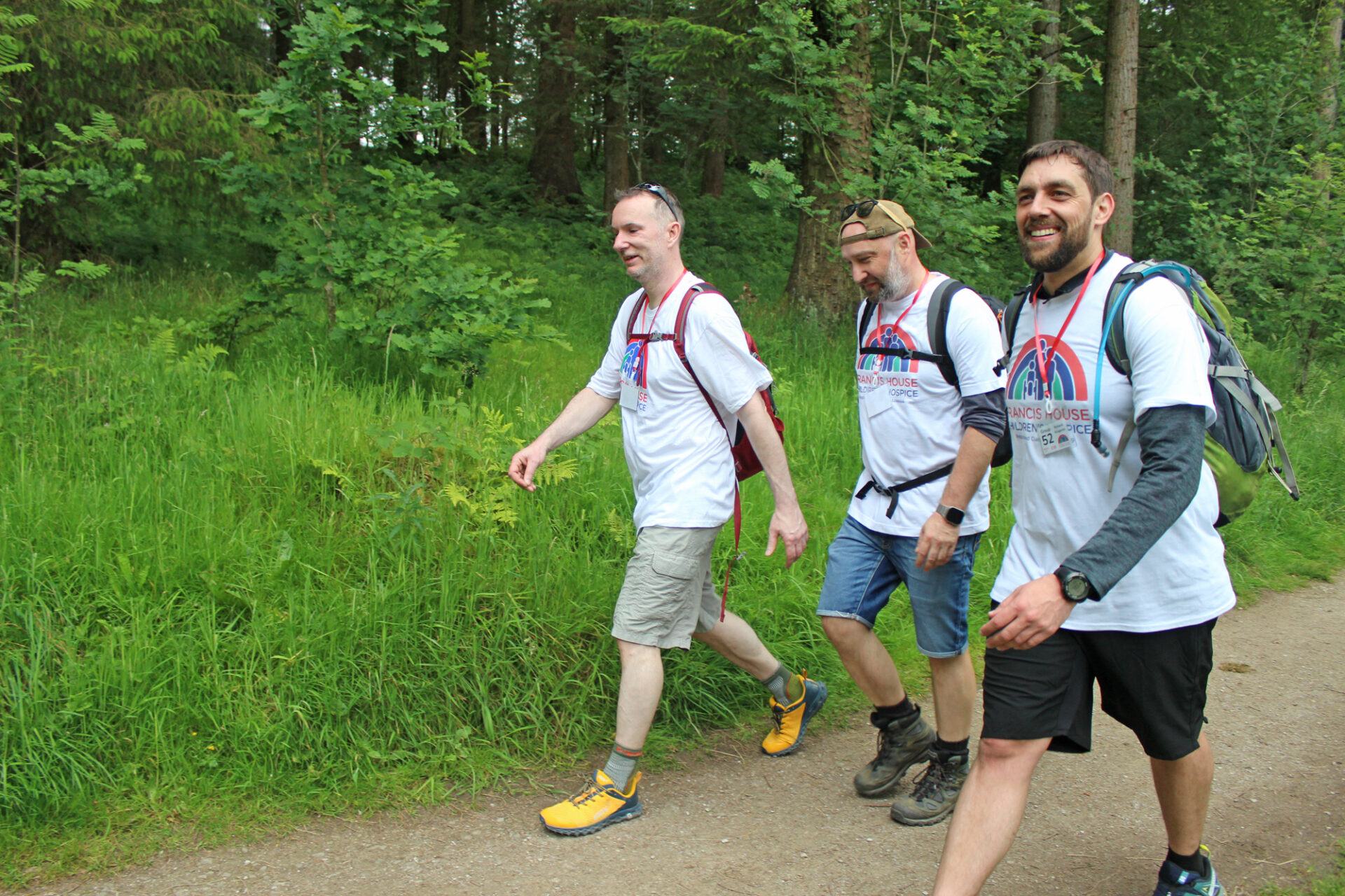 Three men wearing white t-shirts, shorts and walking boots walking along a forest path