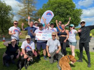 Group of men and women and pet dogs wearing white t-shirts in front of a charity flag and holding a sign saying We did it.