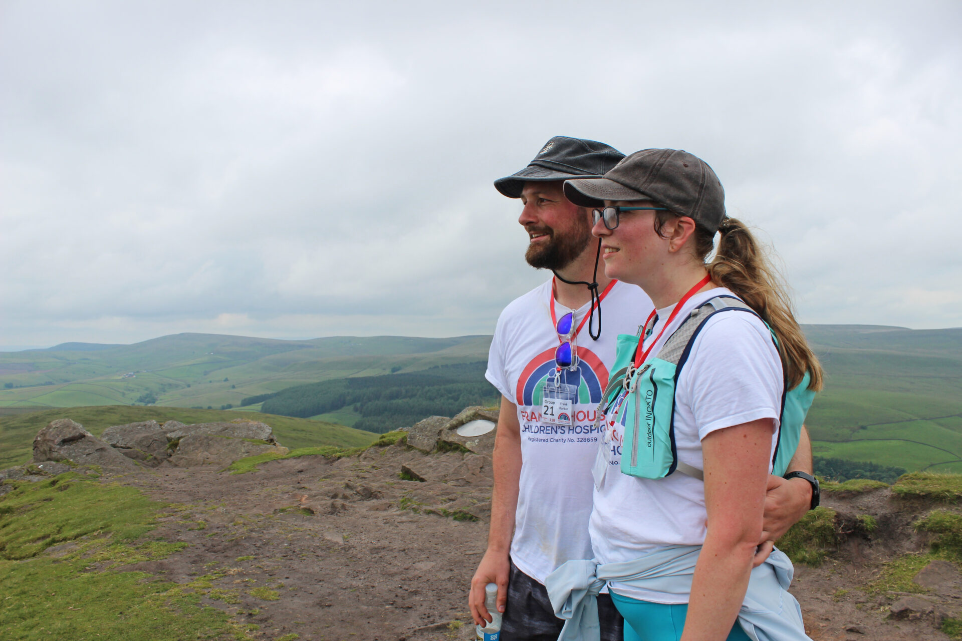 Man and woman wearing shite t-shirts and hats look out at view from a hill