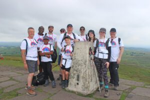 Group of men, women and children wearing white t-shirts and hiking gear stood at the top of Shutlingsloe peak