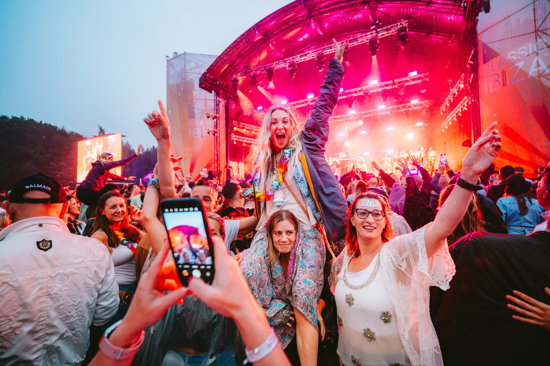 Women in party outfits one holding another on her shoulders in front of a bright pink lit concert stage as another woman takes a photo on a mobile phone