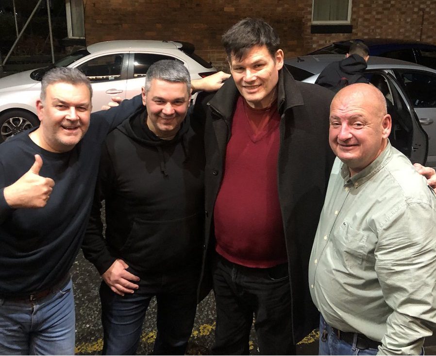 Four men stood in a road one giving a thumbs up smiling at the camera