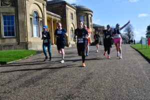 Several participants running past Heaton Park Hall, two of them with arms in the air posing happily for the camera