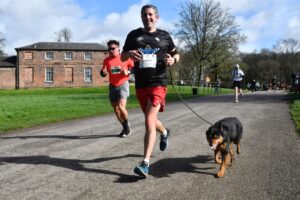 A runner with his dog on a lead head down a tarmac path on the Run Heaton Park course