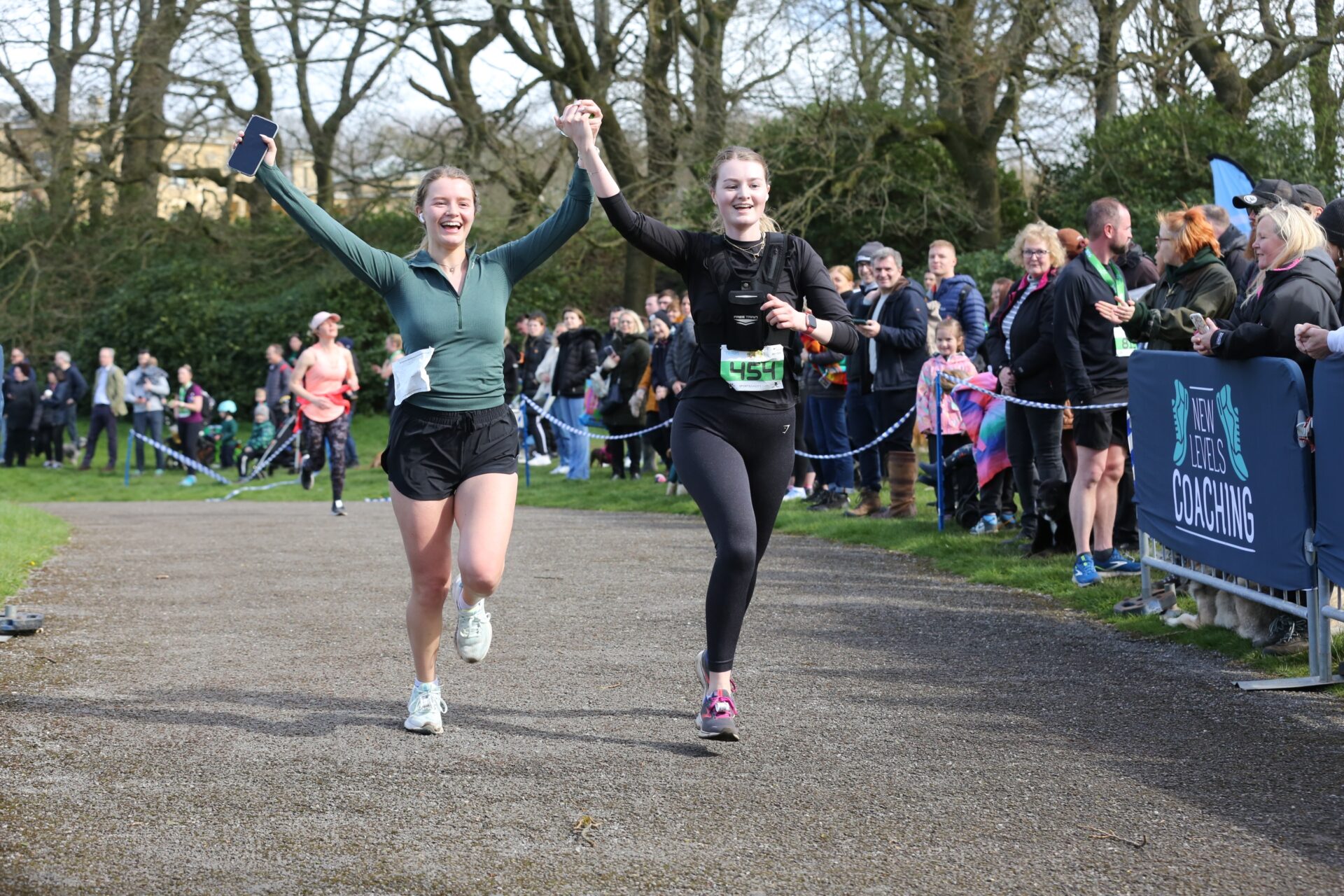 Two cheerful female runners approach the finish line of the Heaton Park run holding their hands in the air in celebration