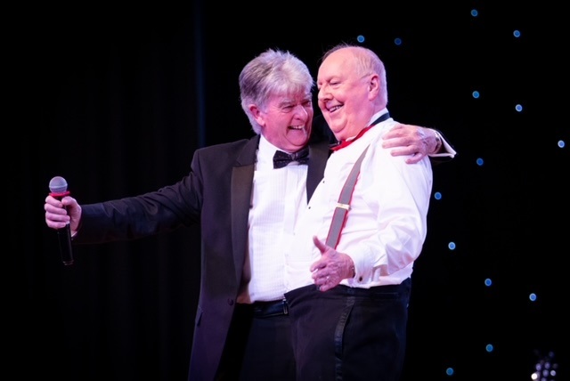 Man wearing a black jacket and tie holding a microphone with his arm around the shoulder of another man stood on a stage