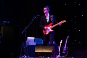 Man dressed as Hank Marvin playing a guitar under a spotlight on a stage