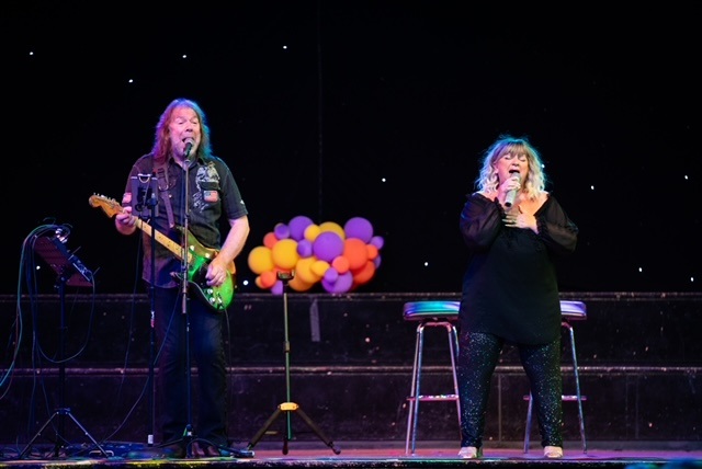 Man playing a guitar stood on a stage with a woman singing in front of coloured balloons