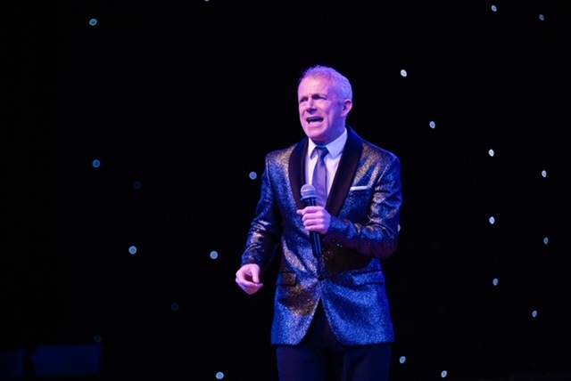 Man dressed in a silver jacket singing under a spotlight on a stage