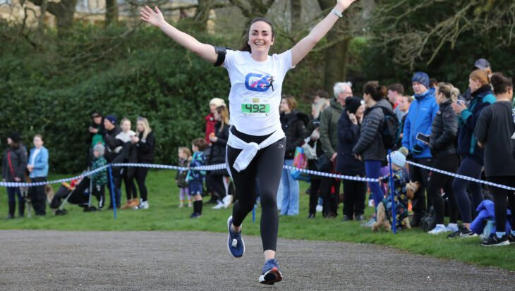 Woman running on a tarmac path in a park with her arms in the air in front of a crowd