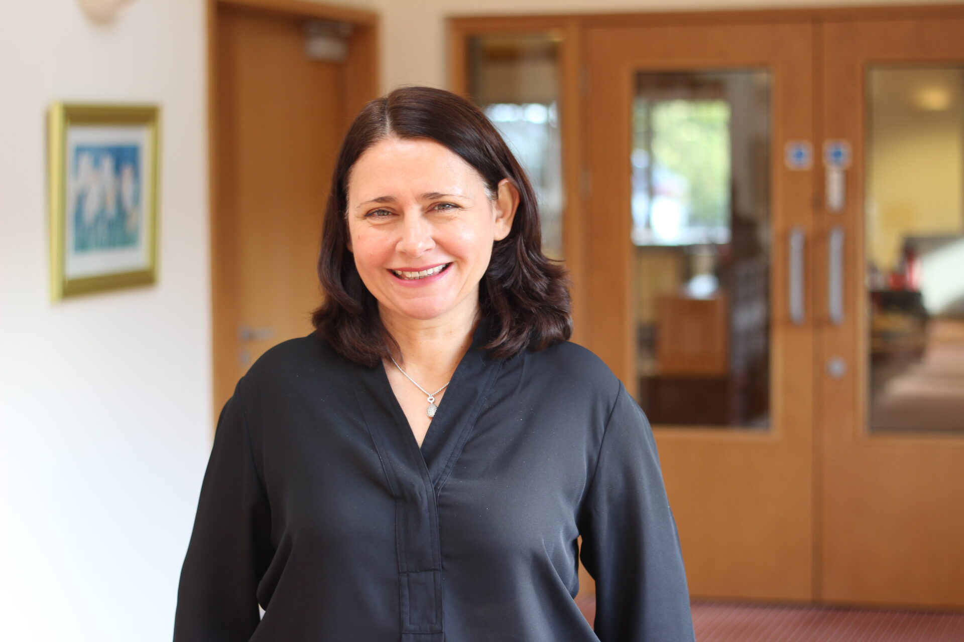 Woman wearing a black shirt stood in a corridor with a picture on the wall and wooden glass doors in the background