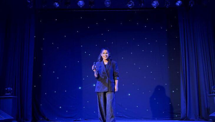 Woman stood on a theatre stage against a blue curtain backdrop holding a microphone