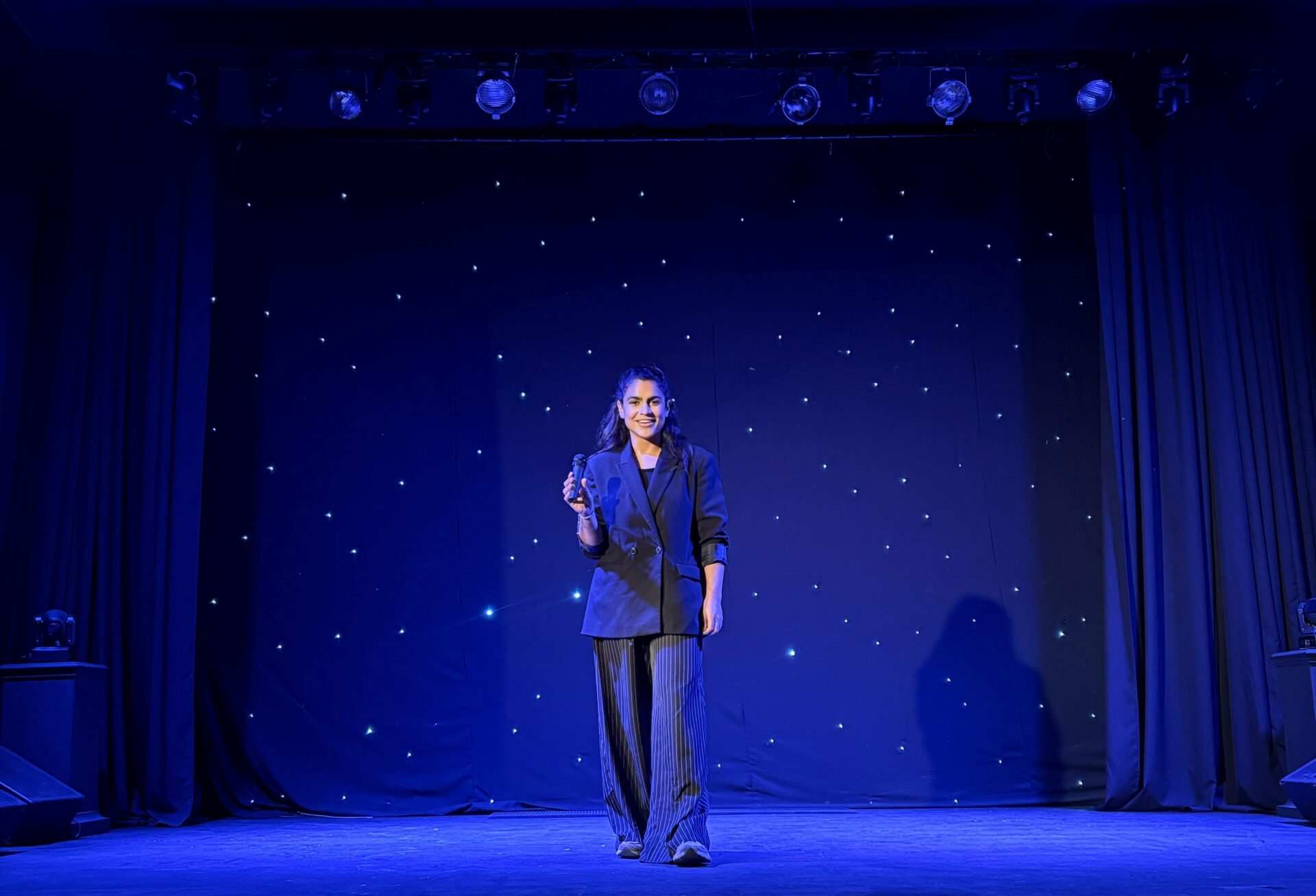 Woman stood on a theatre stage against a blue curtain backdrop holding a microphone